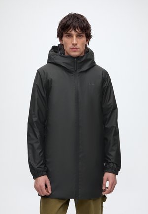 Homme portant une veste imperméable noire à capuche, avec une fermeture éclair intégrale à l'avant et des poignets élastiques, debout devant un fond blanc uni.
