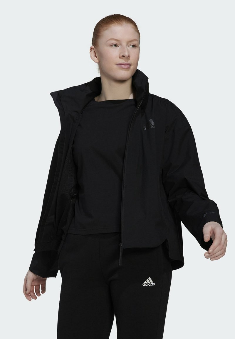 Zwarte jas met hoge kraag, ritssluiting en verstelbare capuchon, gecombineerd met een zwart top en broek. Uitgerust met Adidas-logo accenten.