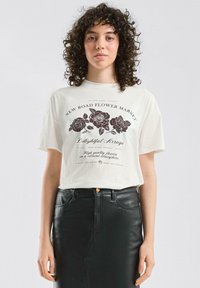 Cremefarbenes T-Shirt mit schwarzem floralen Grafikdesign und Text. Verfügt über kurze Ärmel und eine lockere Passform, kombiniert mit einem hoch taillierten schwarzen Lederrock.