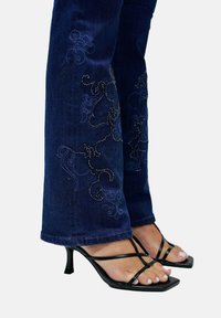 Femme portant un jean brodé bleu foncé et des sandales à talons hauts noires avec des orteils carrés sur un fond blanc.