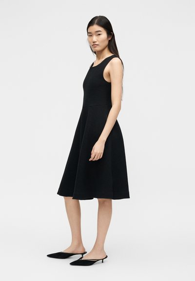 Γυναίκα που φοράει μαύρο αμάνικο φόρεμα μήκους μέχρι το γόνατο με εφαρμοστή μπούστο και φαρδιά φούστα, συνδυασμένο με μαύρα μοκασίνια με χαμηλό τακούνι.