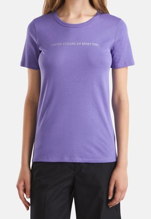 Femme portant un t-shirt violet ajusté à manches courtes avec le texte "UNITED COLORS OF BENETTON." sur la poitrine et un pantalon noir.