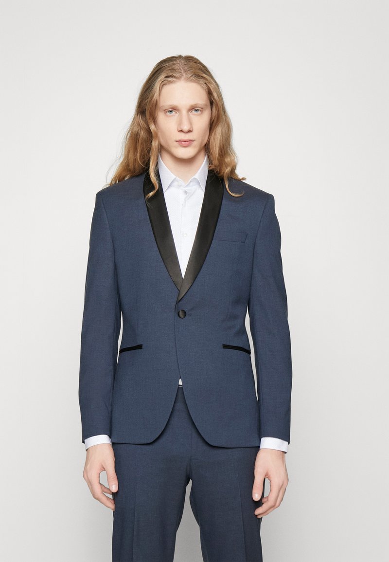 Giacca da completo blu navy con revers in satin nero, chiusura con un solo bottone e due tasche frontali. Abbinata a una camicia azzurro chiaro con colletto.