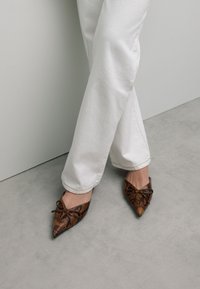 Puntige ballerina's in bruin slangenprint met een gestructureerde afwerking en een decoratieve strikdetail aan de voorkant, gecombineerd met witte broek.