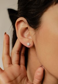 Orecchini a bottone in argento con un design floreale e dettagli in pietre preziose trasparenti, indossati sul lobo dell'orecchio di un modello con una mano che sfiora delicatamente l'orecchio.