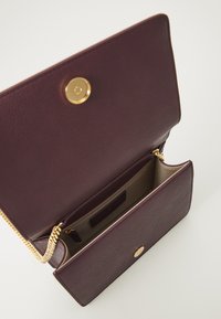 Carteira de couro burgundy com fecho de pressão dourado, apresentando um compartimento interior e um fecho de correia. Textura suave com um padrão de grão sutil.