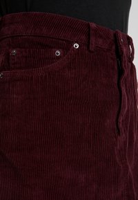 Pantaloni in velluto a coste bordeaux con texture in rilievo, dotati di zip, passanti per cintura e dettagli metallici vicino alle tasche.