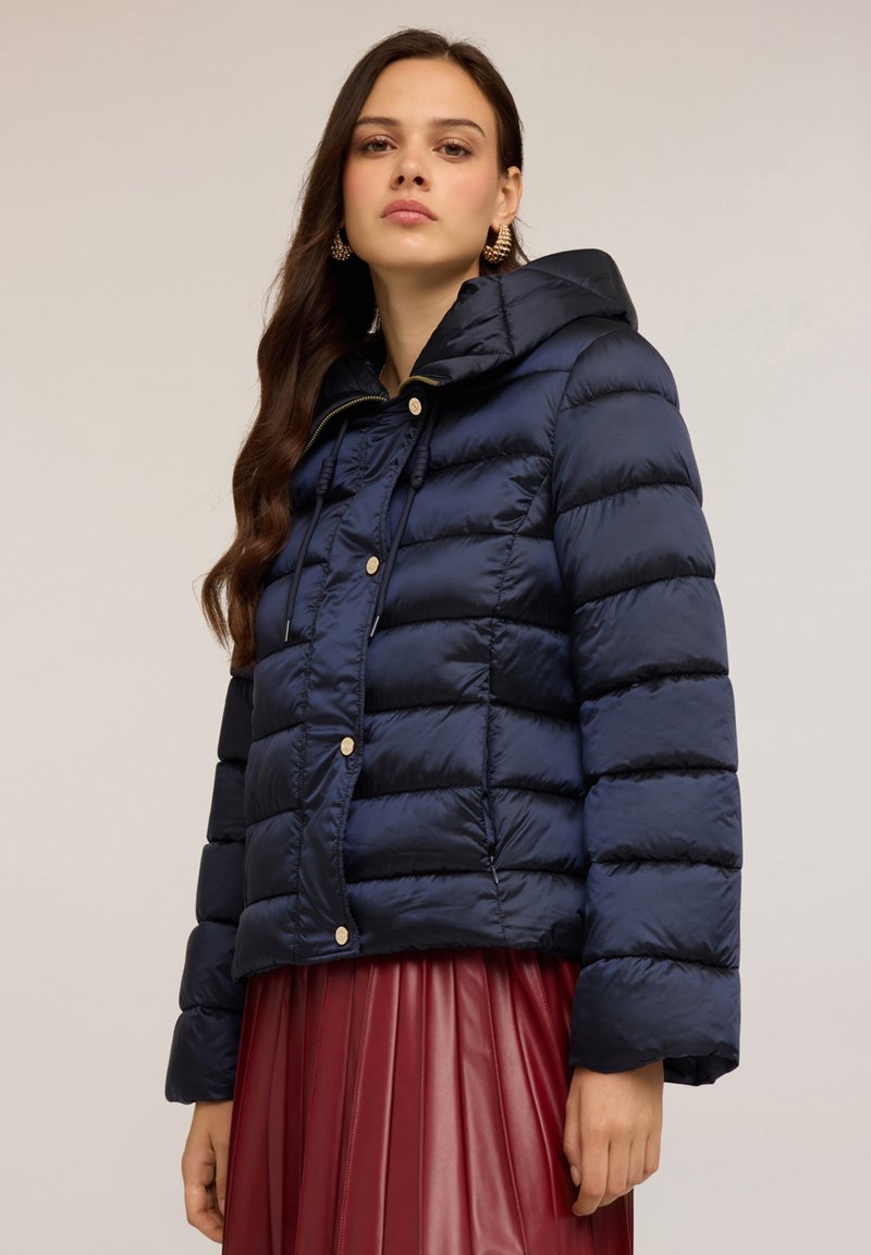 Jachetă puffer bleumarin cu glugă, având cusături orizontale și închidere cu nasturi. Textură netedă și design ușor supradimensionat.