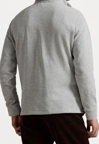 Pull-over gris à manches longues avec tissu texturé, coupe décontractée et léger col, associé à un pantalon marron foncé.