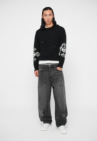 Un modèle masculin porte un pull noir avec des motifs de crâne sur les manches, associé à un jean gris foncé ample et des baskets blanches, se tenant devant un fond blanc.