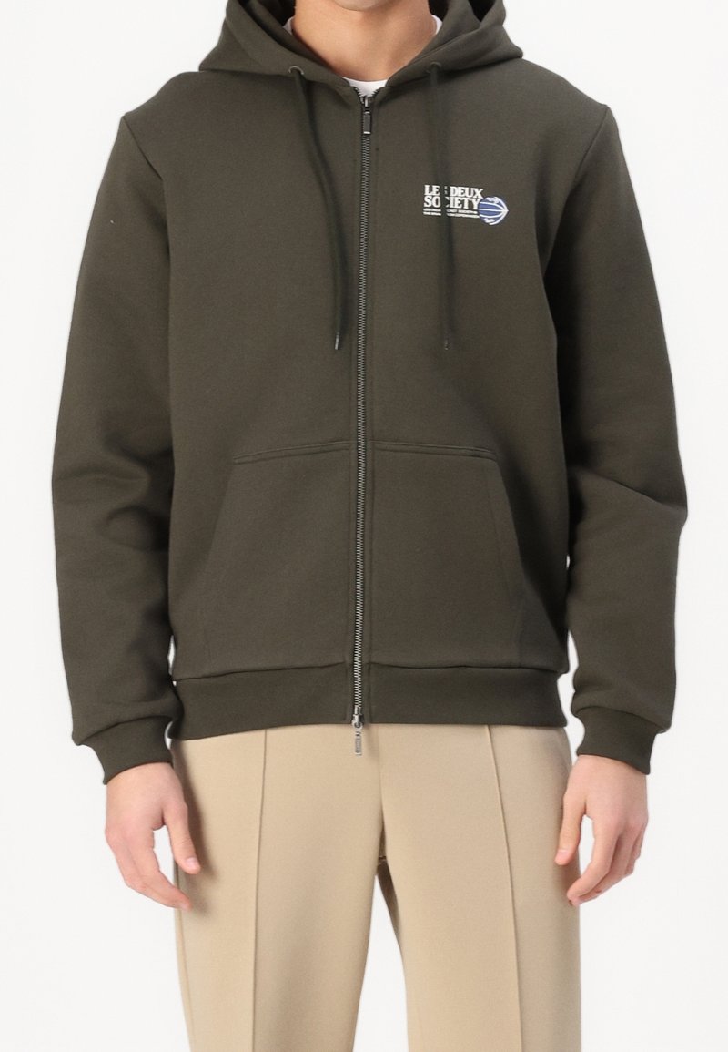 Homme portant un sweat à capuche vert zippé avec un petit logo blanc et bleu sur la poitrine gauche et un pantalon beige sur fond blanc.