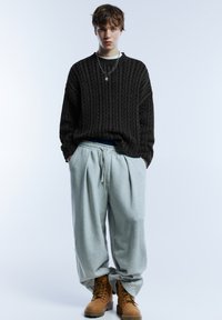 Čierny pletený sveter s textúrovaným vzorom, svetlošedé tepláky so sťahovacou šnúrkou a hnedé šnurovacie topánky.