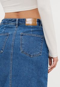 Denim midi suknja u plavoj boji s uskim strukom, stražnjim džepovima i smešnim kožnim etiketom. Ima ravni kroj i teksturiranu tkaninu.