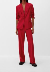 Costume rouge sur mesure avec un blazer ajusté à revers et un pantalon à jambes larges. Tissu lisse avec une texture légèrement brillante. Chaussures noires.