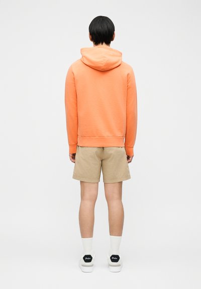 Άτομο που φοράει πορτοκαλί φούτερ με κουκούλα, μπεζ σορτς, λευκές κάλτσες και λευκά παπούτσια Polo, στέκεται με την πλάτη γυρισμένη σε απλό φόντο.