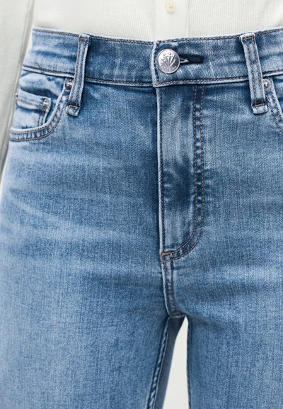 Μπλε τζιν παντελόνι με κλείσιμο με κουμπιά μπροστά, μπροστινές τσέπες και ορατή ραφή, φορεμένο με λευκή ριμπ πλεκτή μπλούζα.