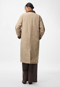 Μακρύ μπεζ παλτό με καφέ γιακά, διαθέτει ευθεία γραμμή, λεία υφή και σχισμή στην πλάτη. Συνδυασμένο με φαρδιά καφέ παντελόνια.