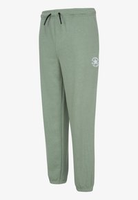 Grønne joggebukser laget av mykt stoff, med snøring i livet, smale ben og en hvit logo på venstre lår.