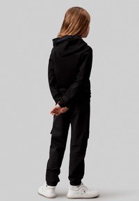 Felpa nera con un logo sul collo, abbinata a pantaloni cargo neri e sneakers bianche. Texture liscia e vestibilità aderente.