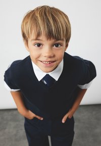 Uśmiechnięty chłopiec z jasnobrązowymi włosami, ubrany w granatowy sweter, białą koszulę i granatowy krawat, stojący z rękami w kieszeniach, zwrócony w stronę kamery.