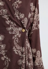 Wijnrode wikkeljurk met crème bloemen-paisleypatroon, gestructureerde stof en een decoratieve gouden knoop in de taille.