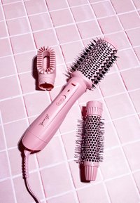 Brosse à cheveux en céramique ronde avec un manche en plastique rose et des têtes de brosse détachables, dotées de poils noirs ; posée sur une surface carrelée rose clair.