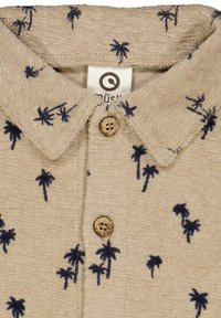 Camicia beige con colletto, decorata con ricamo di palme blu navy, dotata di bottoni in legno e una finitura texture. Etichetta visibile sotto il colletto.