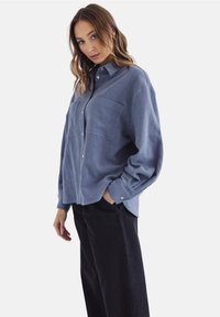Kobieta z jasnobrązowymi włosami, ubrana w luźną niebieską koszulę z guzikami i czarne spodnie, stojąca z jedną ręką w kieszeni na białym tle.