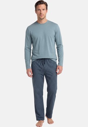Man staat blootsvoets, draagt een lichtblauw shirt met lange mouwen en donkerblauwe pyjamabroek met patroon tegen een effen achtergrond.