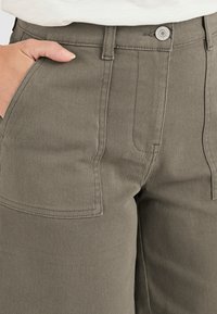 Pantaloni in misto cotone verde oliva con vita alta, dotati di tasche frontali e chiusura con bottone metallico. Tessuto strutturato con cuciture visibili.