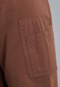 Vêtement en coton marron avec une poche latérale dotée de coutures horizontales. Une petite étiquette noire est fixée près de la poche. Tissu texturé.