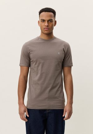 Ung mand set forfra iført en taupe kortærmet T-shirt med lille "II" logo på brystet og mørkeblå jeans mod en ensfarvet baggrund.