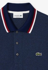 Polo Lacoste bleu marine avec col rayé rouge, blanc et noir, trois boutons et logo crocodile vert sur la poitrine.