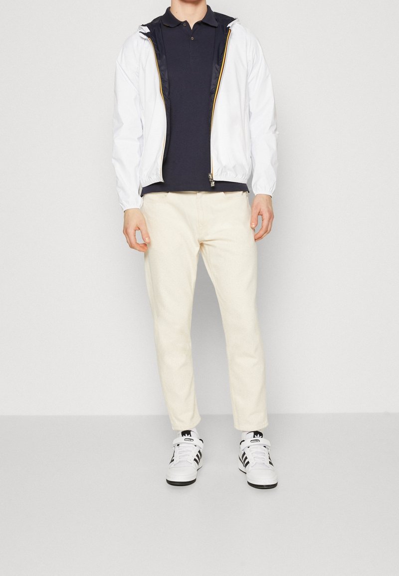 Personne debout portant une veste blanche zippée sur un polo noir, un pantalon couleur crème et des baskets blanches Adidas avec des rayures noires.