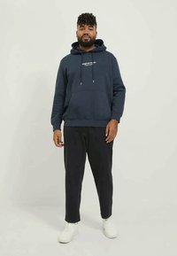 Man with beard wearing navy hoodie, black pants, and white sneakers standing against plain light background.