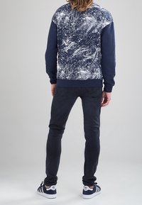 Sudadera azul marino con un diseño de manchas de pintura blanca en la parte superior de la espalda, combinada con jeans oscuros y zapatillas negras y blancas.