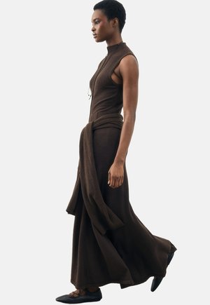 Vrouw loopt en draagt een donkerbruine mouwloze maxi-jurk met een bijpassende trui om de taille gestrikt en zwarte ballerina's.
