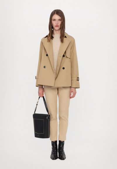 Manteau trench beige à double boutonnage avec des boutons noirs, associé à un pull beige et un pantalon. Sac seau noir avec des accents dorés et des bottes cloutées.