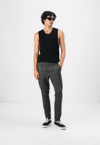 Schwarzes ärmelloses Oberteil, kombiniert mit grauen karierten Hosen. Die Hosen haben einen Knopfverschluss und werden durch schwarze Sneakers ergänzt.