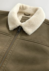 Olivgrüne Kunstlederjacke mit einem cremefarbenen, flauschigen Kragen und einem Reißverschluss, ausgestattet mit genähten Details an den Schultern und Seiten.