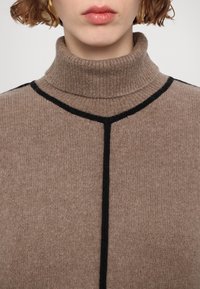 Personne portant un pull à col roulé côtelé marron avec des lignes noires formant un T sur le devant, visible du cou à la poitrine.