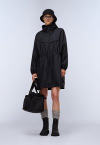 Giacca impermeabile nera con vita elastica, chiusura zip, grandi tasche, abbinata a un cappello a secchiello nero, calze grigie e stivali alla caviglia.