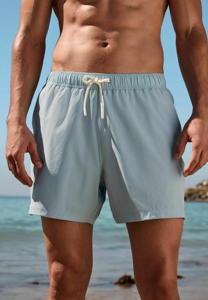 Hemdloser Mann trägt hellblaue Badeshorts mit Kordelzug und steht am Meer vor klarem Himmel.