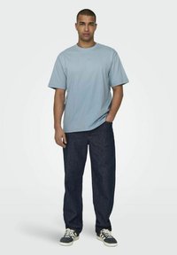 Helleblaue Kurzarm-T-Shirt kombiniert mit dunkelblauen Jeans. Das Modell steht lässig mit einer Hand in der Tasche und trägt schwarz-weiße Sneakers.