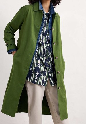 Femme portant un long manteau vert sur une chemise bleue à motifs et un pantalon beige, debout les mains dans les poches du manteau devant un fond uni.