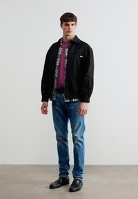 Νεαρός άνδρας που στέκεται μπροστά σε απλή λευκή τοίχο, φοράει μαύρο μπουφάν, πουκάμισο καρώ, μοβ μπλουζάκι, μπλε τζιν και μαύρα παπούτσια, κοιτάζοντας ελαφρώς προς τα δεξιά.