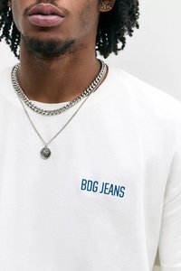 Artimas kadras asmens, dėvinčio balta BDG Jeans marškinius, dvi sidabro grandineles ir mažus auskarus su rožiniais akcentais.