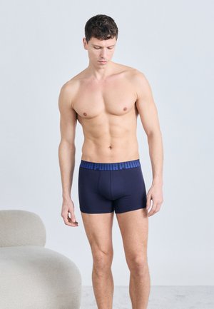 Homme torse nu portant un boxer bleu marine avec une ceinture élastique à lettres bleues, debout à côté d'une chaise arrondie gris clair contre un mur clair uni.