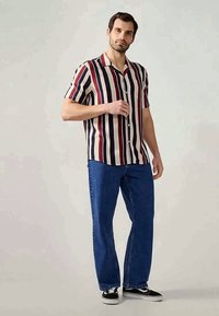 Chemise à manches courtes avec des rayures verticales rouges et marines sur un fond crème, associée à un jean en denim bleu et des baskets noires.