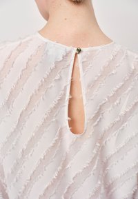 Weiße Bluse aus strukturiertem Stoff mit ausgefransten horizontalen Streifen, einem weiteren Ausschnitt am Rücken und einem kleinen Knopfverschluss.
